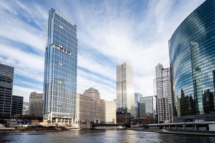 Modern skyscrapers with sleek curtainwall facades line the Chicago River under a partly cloudy sky.